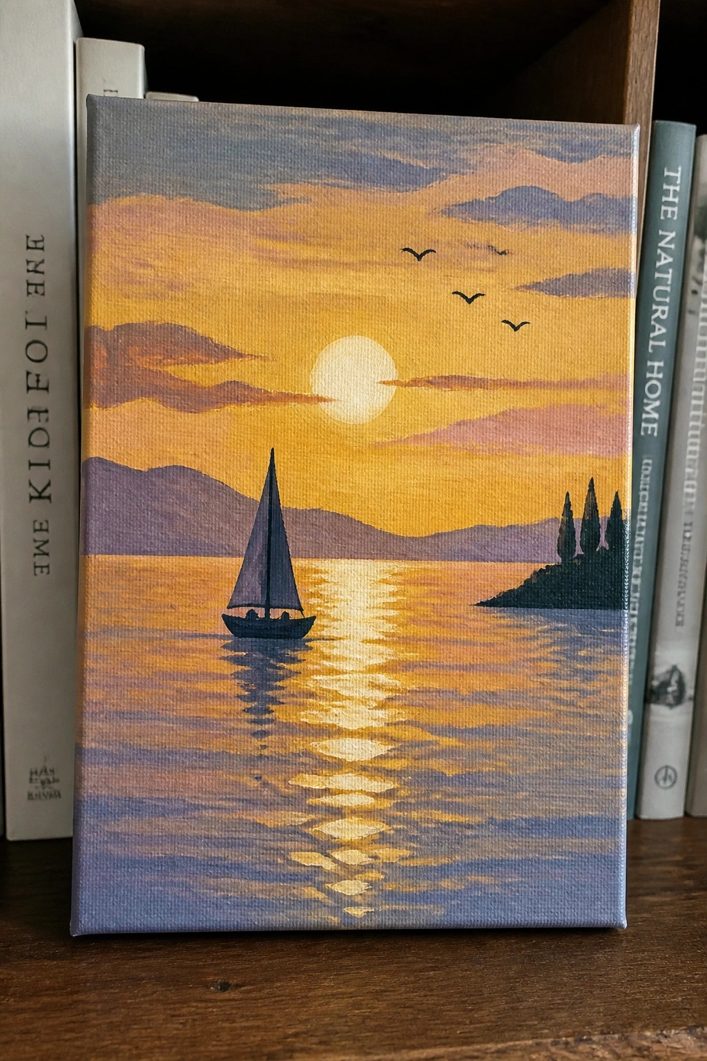 A canvas painting of a sailboat on a lake at sunset with golden reflections, mountains, birds, and a small island, placed between books on a shelf.
