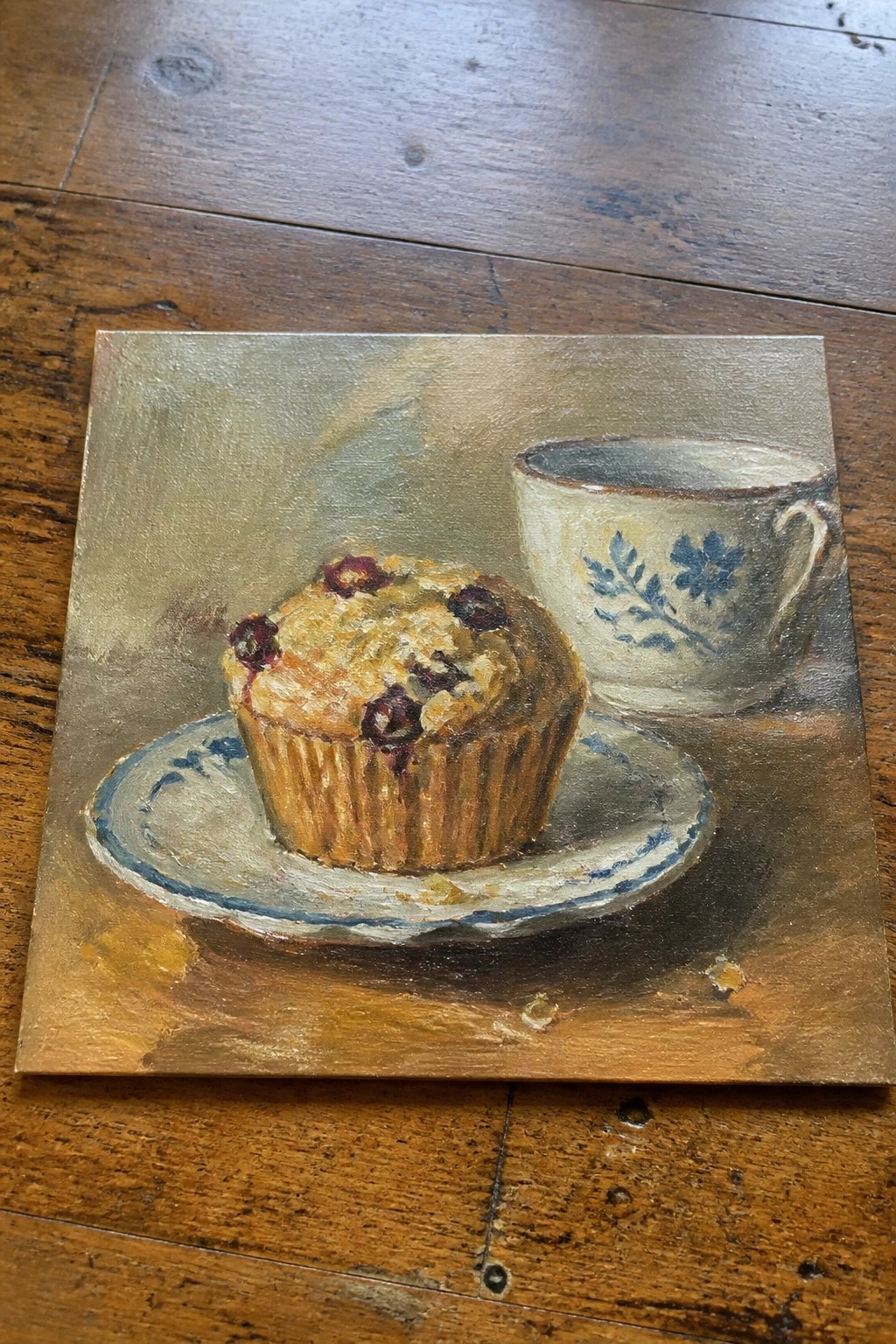 Realistic painting of blueberry muffin on plate beside blue floral teacup on wood.