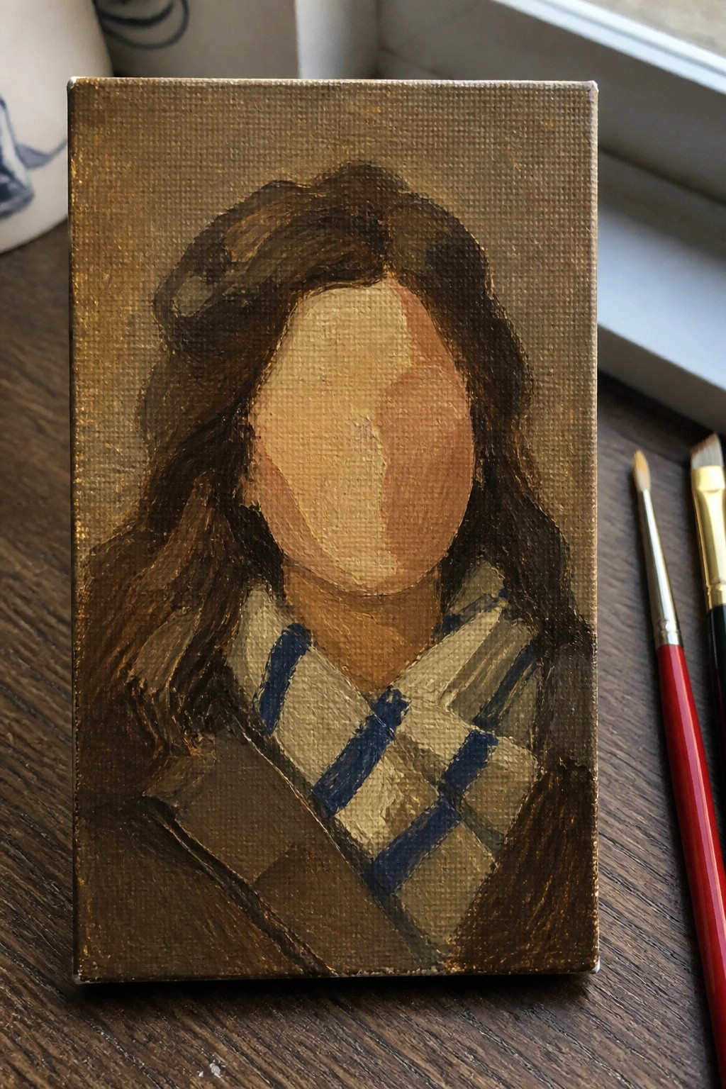 Small canvas portrait with dark wavy hair, blank face, and plaid scarf beside paintbrushes.