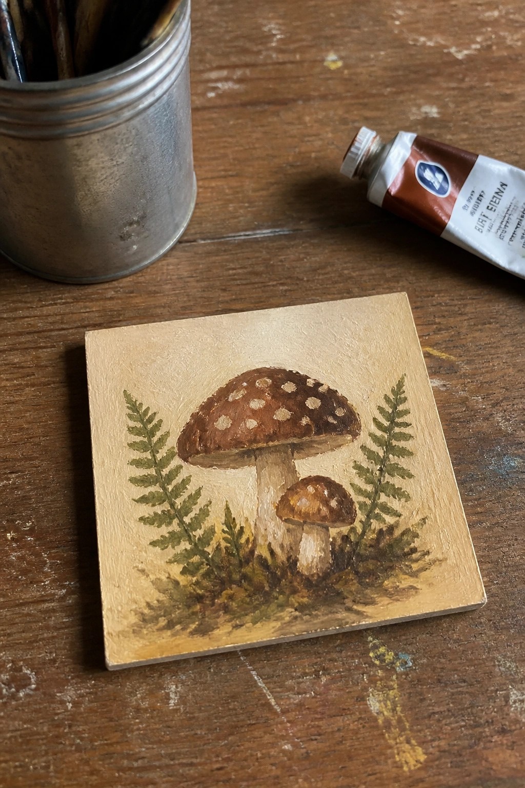 Small square painting of two spotted mushrooms with ferns beside burnt sienna tube and brushes.