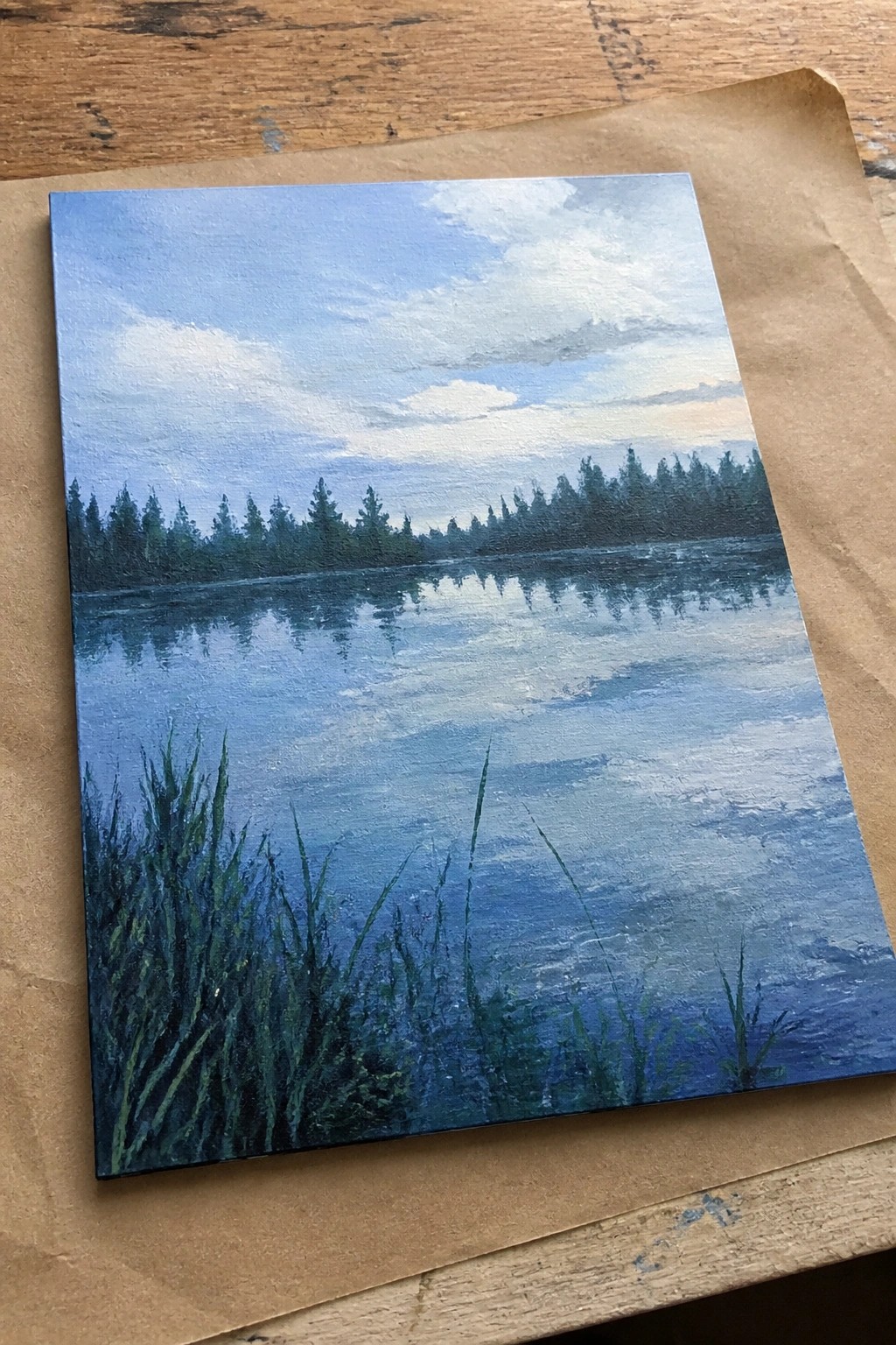 Painting of calm blue lake with pine tree reflections and foreground reeds on wood