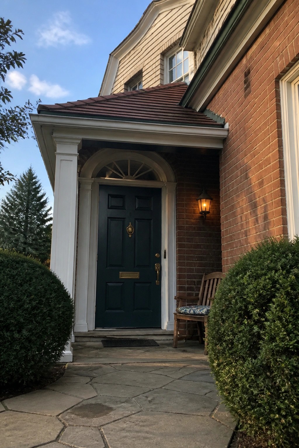 Brick house porch with dark green door, white columns, wooden chair and shrubs
