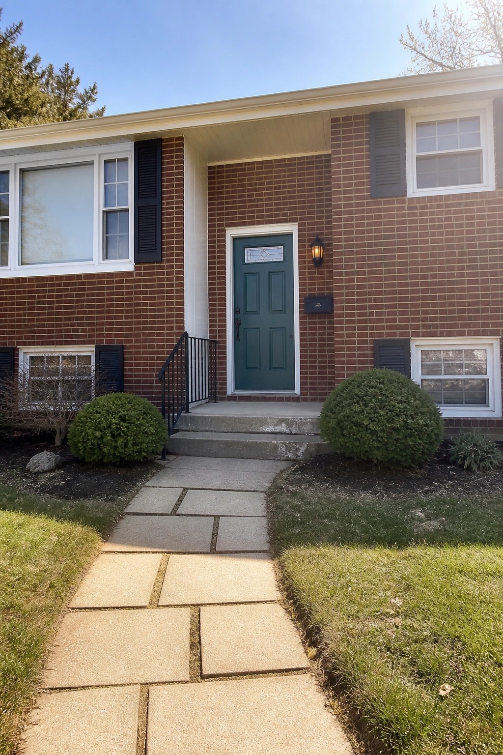 Brick house with teal door, concrete steps, stone walkway, black shutters, and bushes.