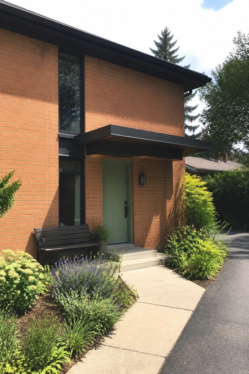 Modern brick house with green door, black awning, bench, and garden.