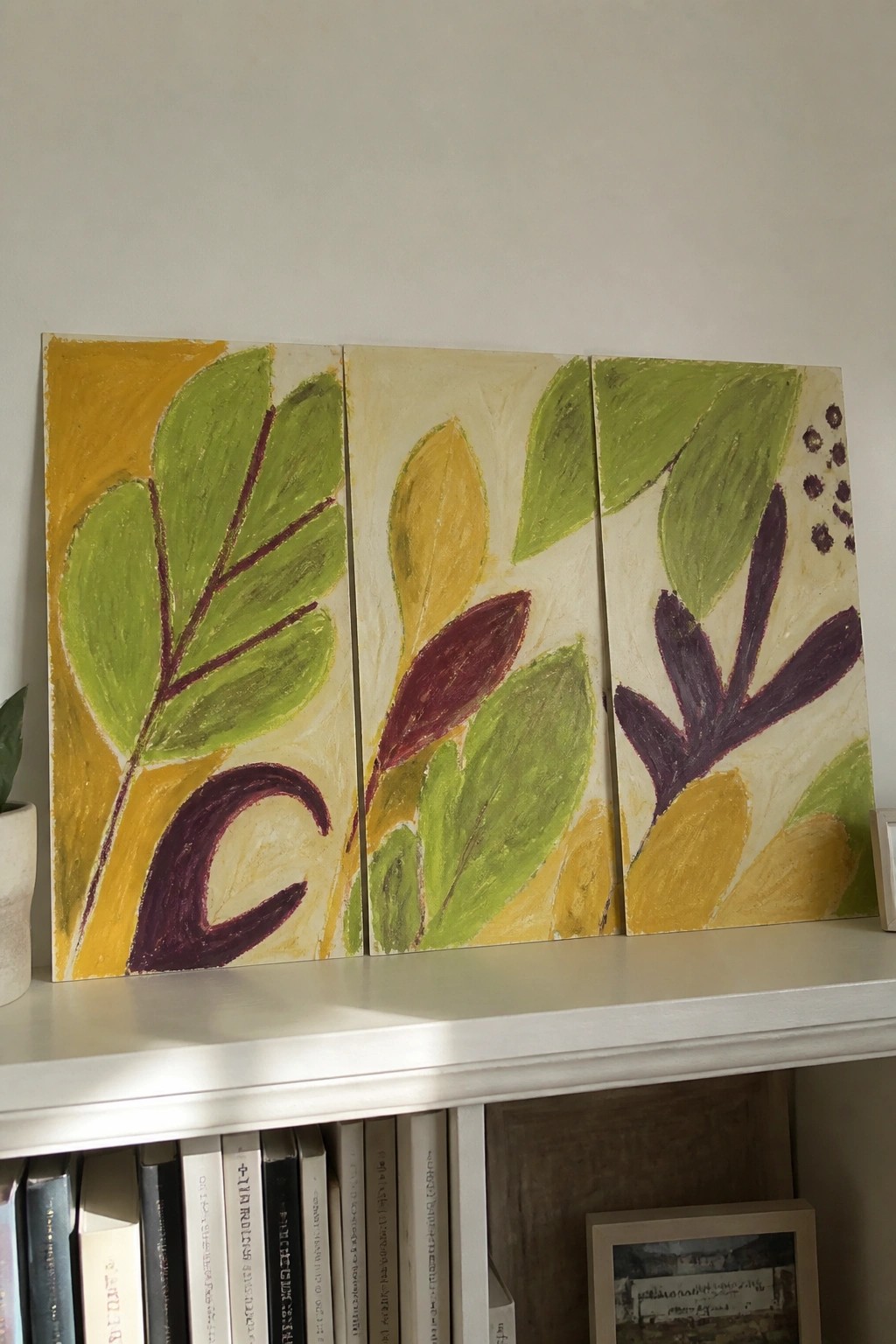 Abstract triptych of green, yellow, and purple leaves on a white shelf above books