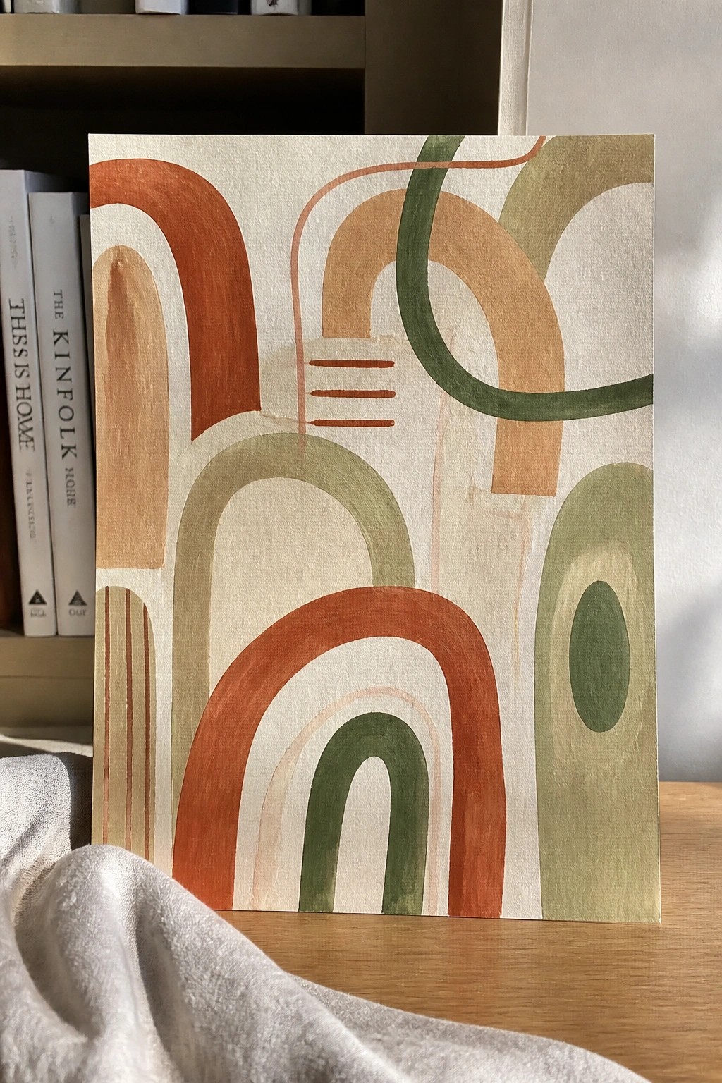 Abstract artwork with terracotta and green arches on wooden table beside books and fabric.