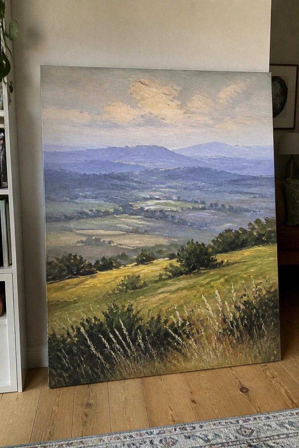 Large landscape painting of green fields and distant blue mountains on wooden floor