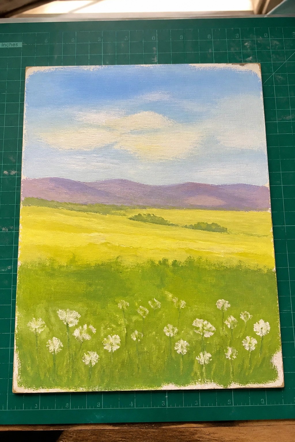Pastoral landscape painting of yellow fields, purple mountains, and white flowers on green mat