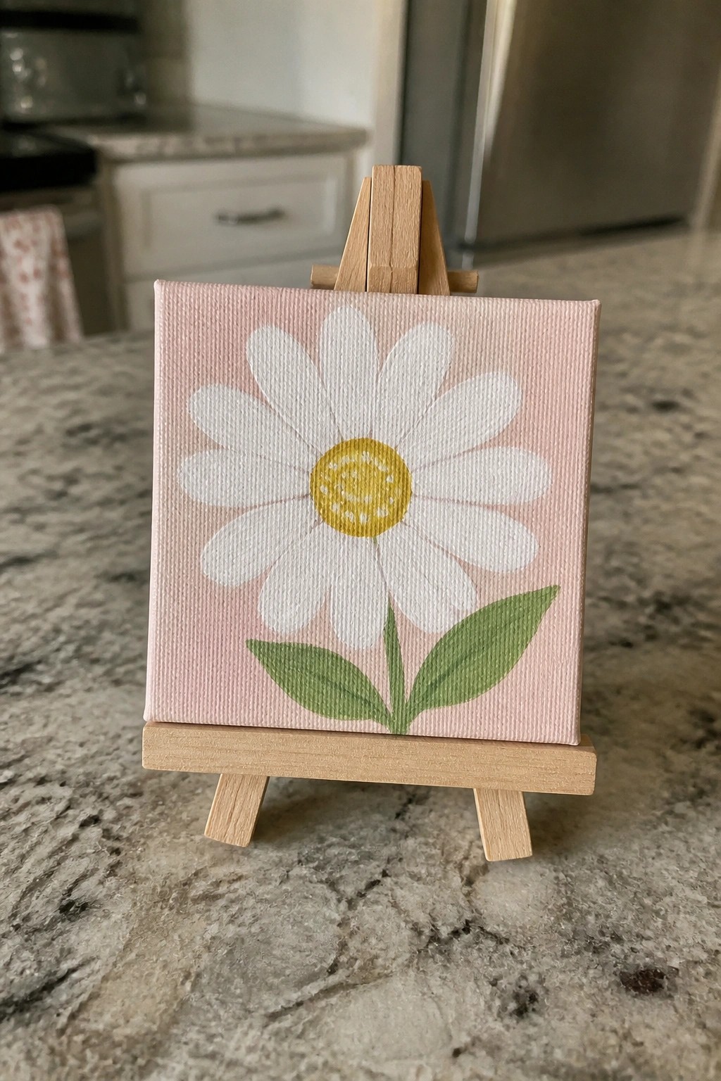 Small pink canvas with white daisy on wooden easel atop marble counter