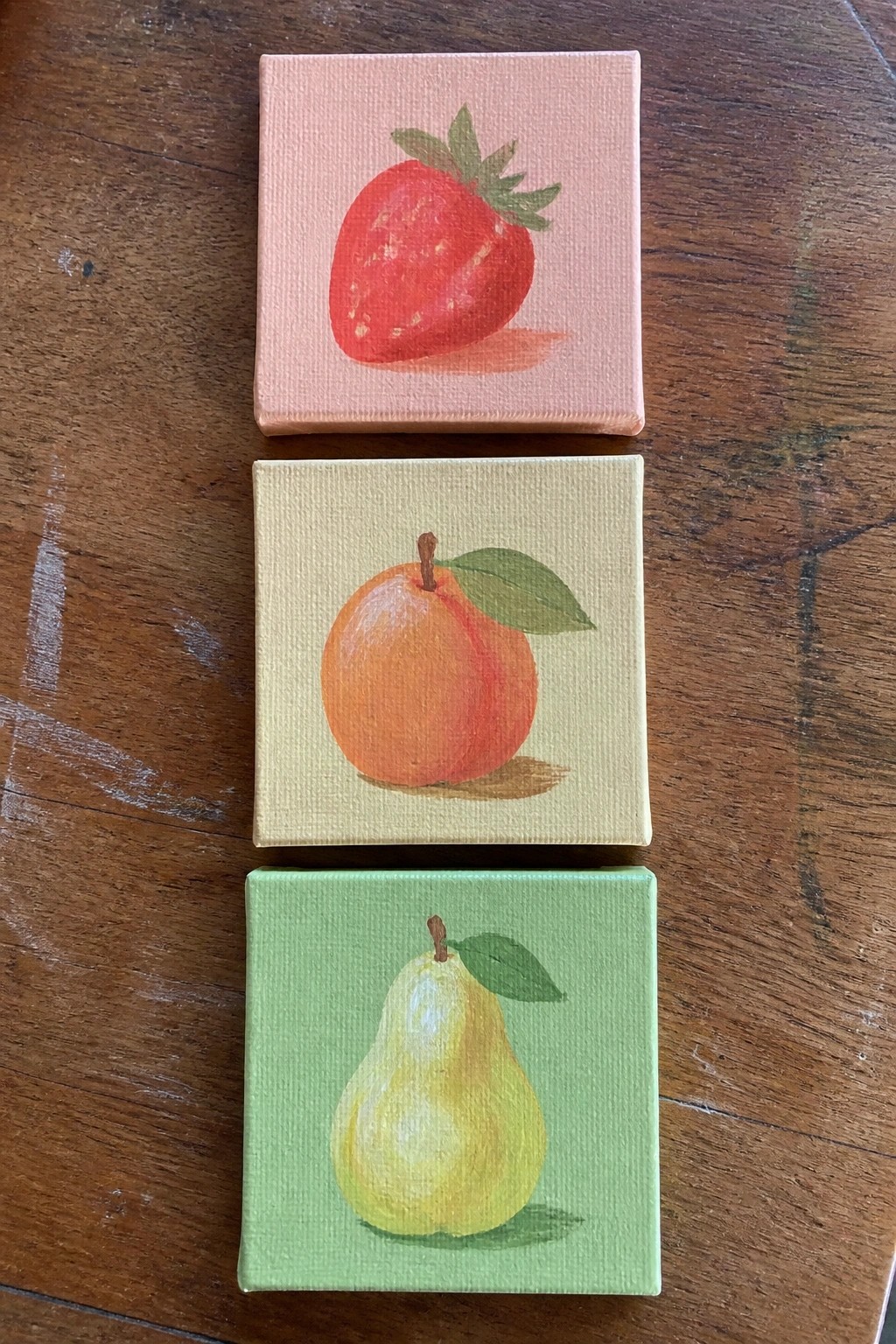 Three small canvases with strawberry on pink, peach on yellow, and pear on green.