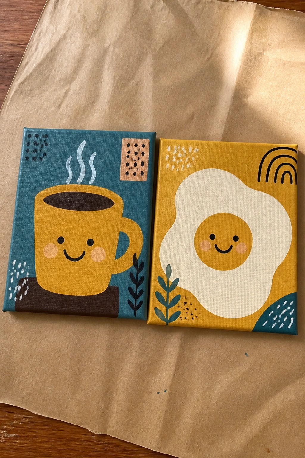 Two small canvases side by side on brown paper showing a cute coffee mug painting and a cute fried egg painting.