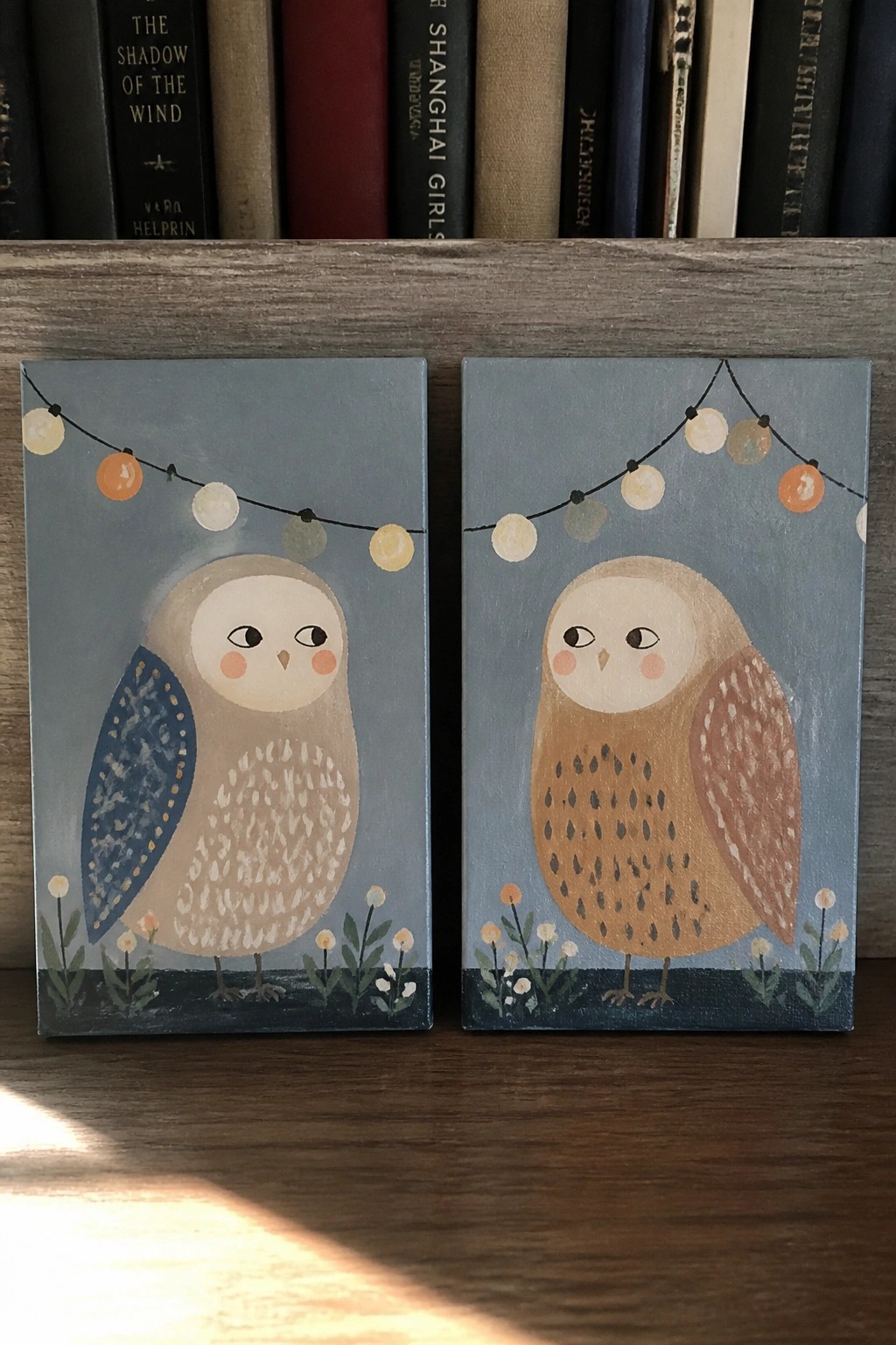 Two whimsical owl paintings with string lights and flowers before books on wood.