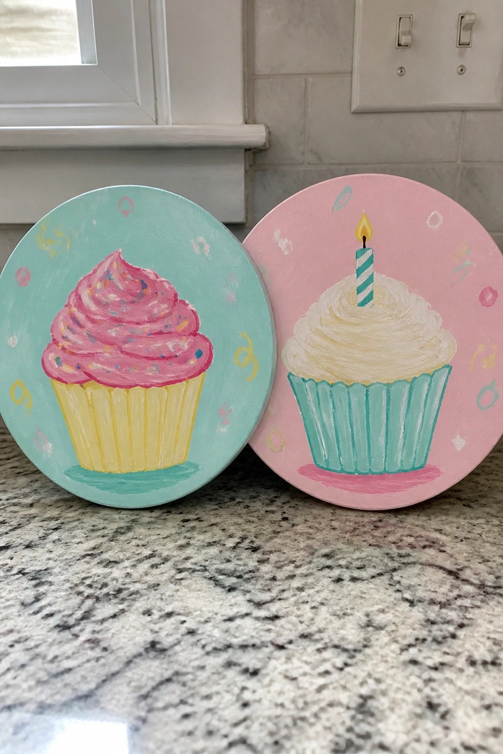 Two round cupcake paintings on a granite counter, one pink-frosted on teal and one white-frosted with a candle on pink.