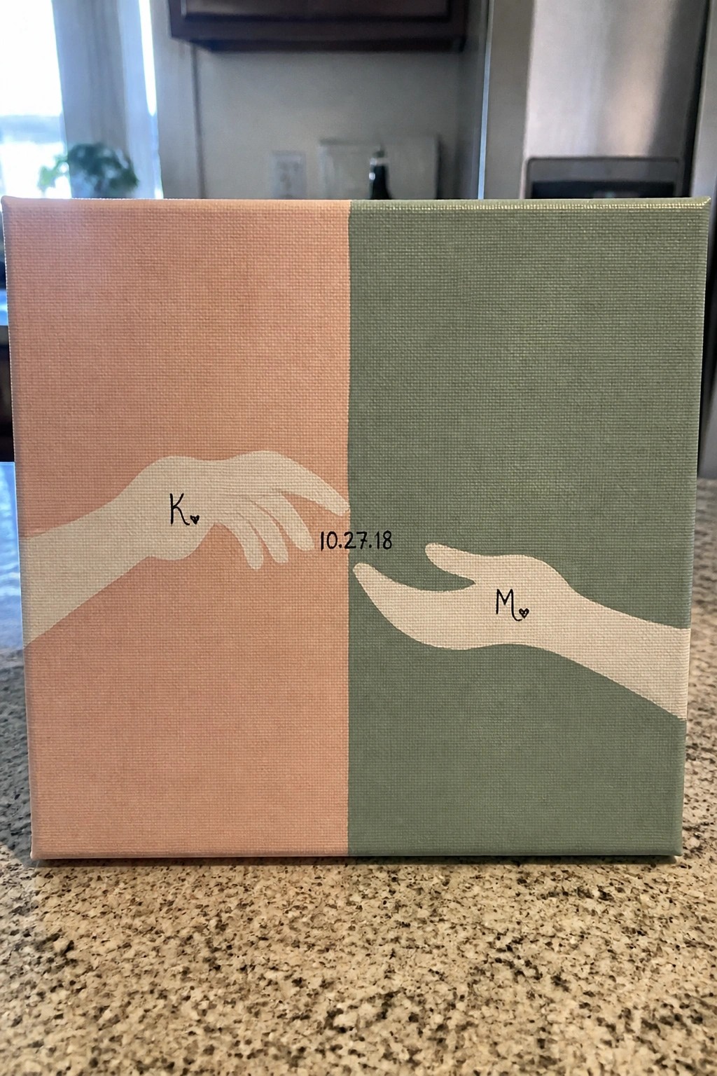 A square canvas split vertically into peach and green sections with two hands reaching toward each other, initials K and M, and the date 10.27.18 in the center.