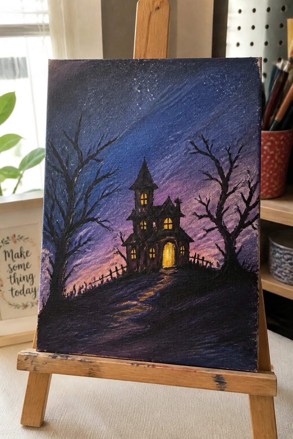 Haunted house painting with glowing windows, bare trees, and starry purple sky on easel