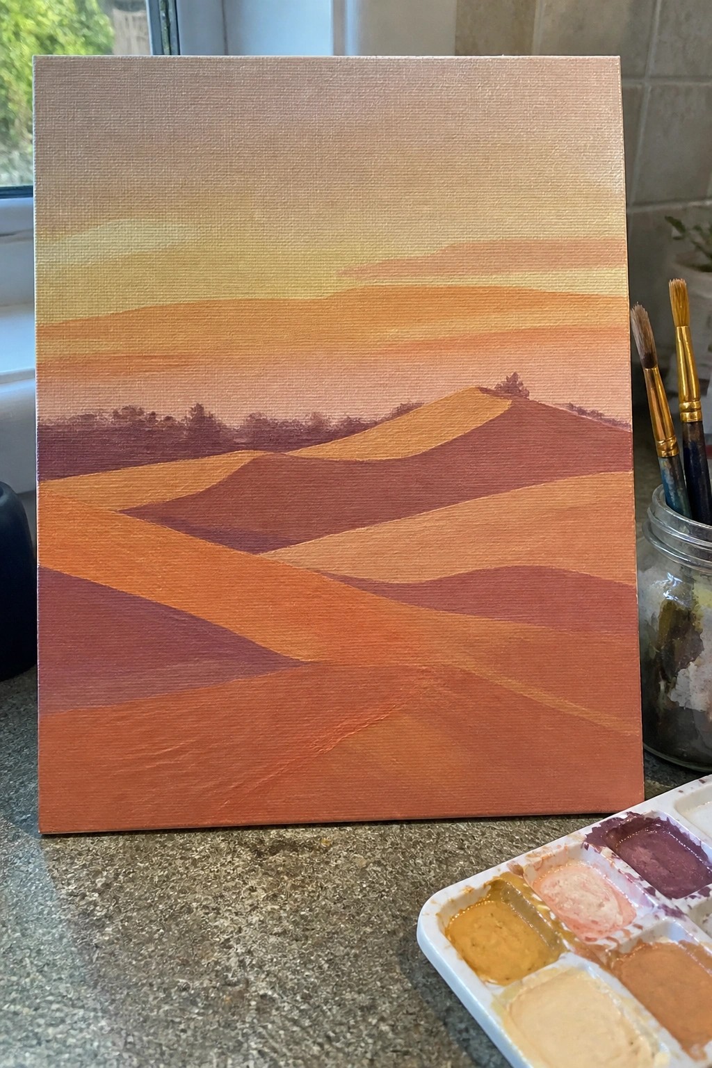 Canvas with abstract sunset landscape in reds and oranges beside paint palette and brushes
