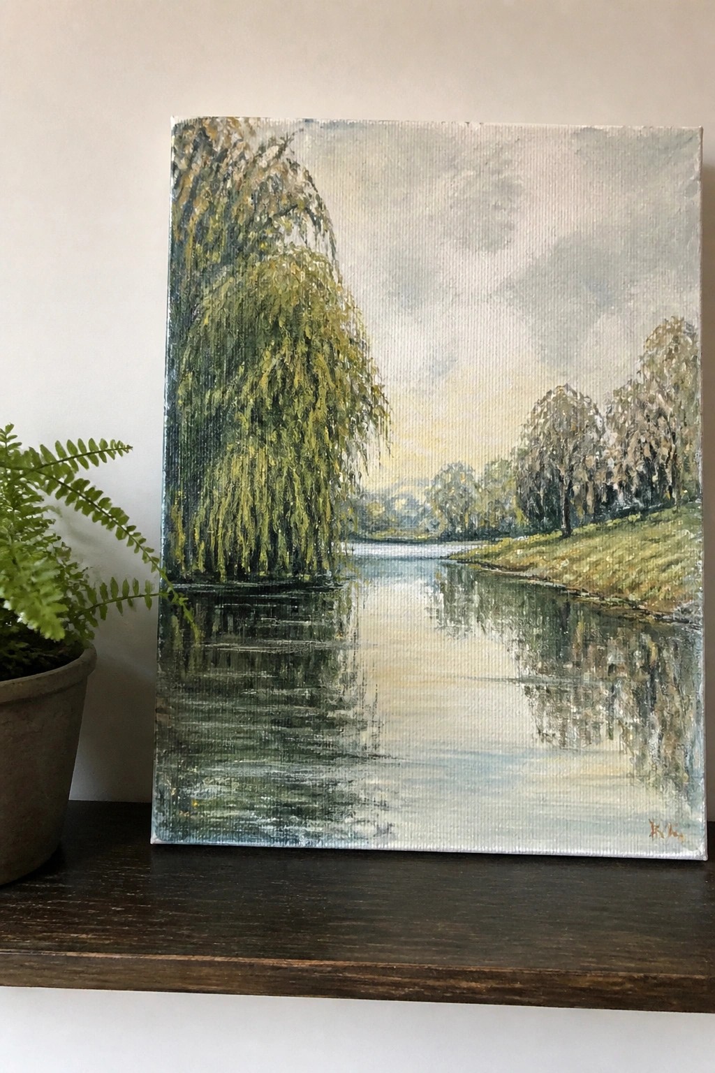 River landscape painting with weeping willow reflections beside a potted fern plant.