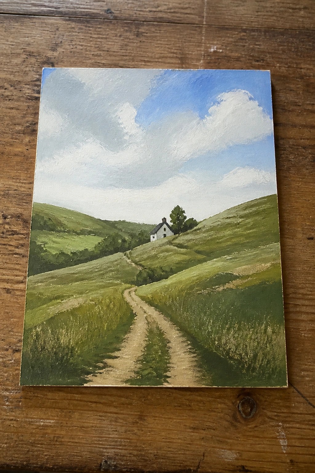 Winding dirt path leads through rolling green hills to a white house under blue sky.