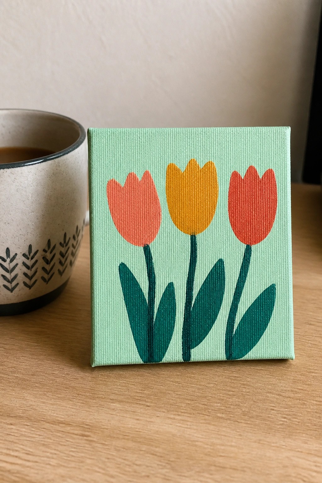 Small canvas with orange, yellow, and red tulips on mint green beside a mug.