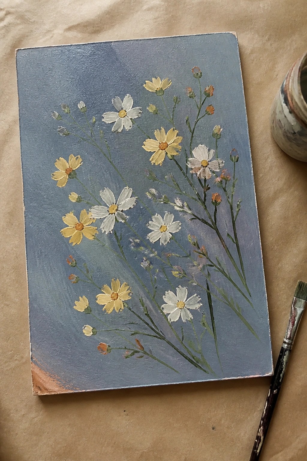 Painting of white and yellow flowers on blue canvas with brush and jar nearby