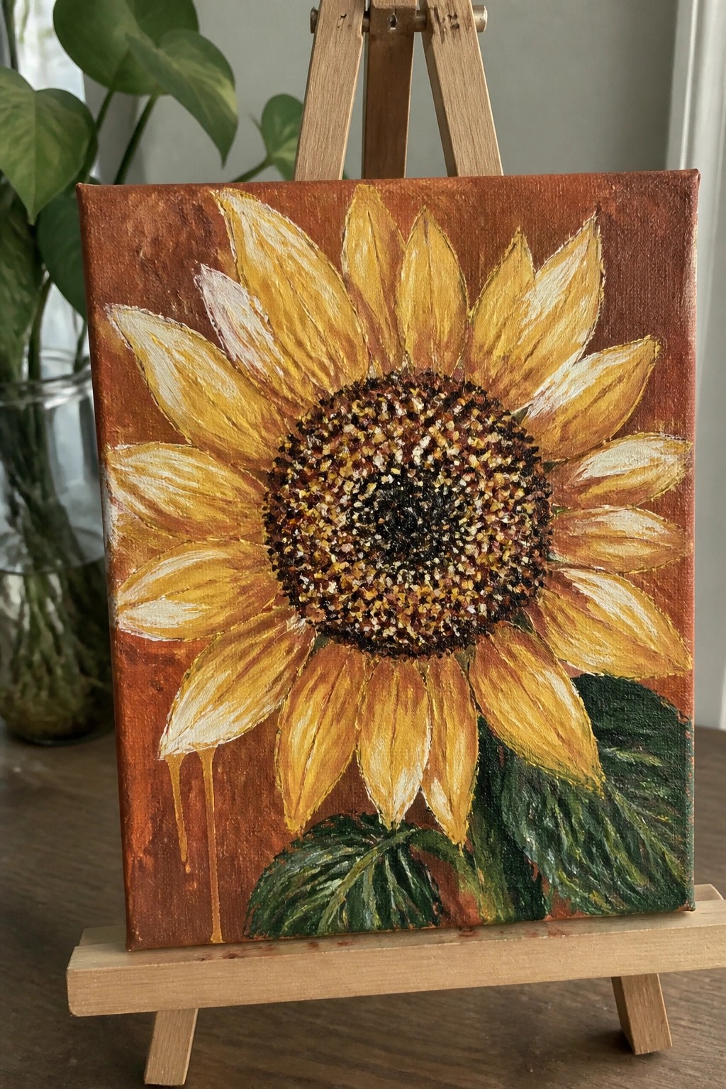 Sunflower painting with yellow petals, brown center, and green leaves on wooden easel.