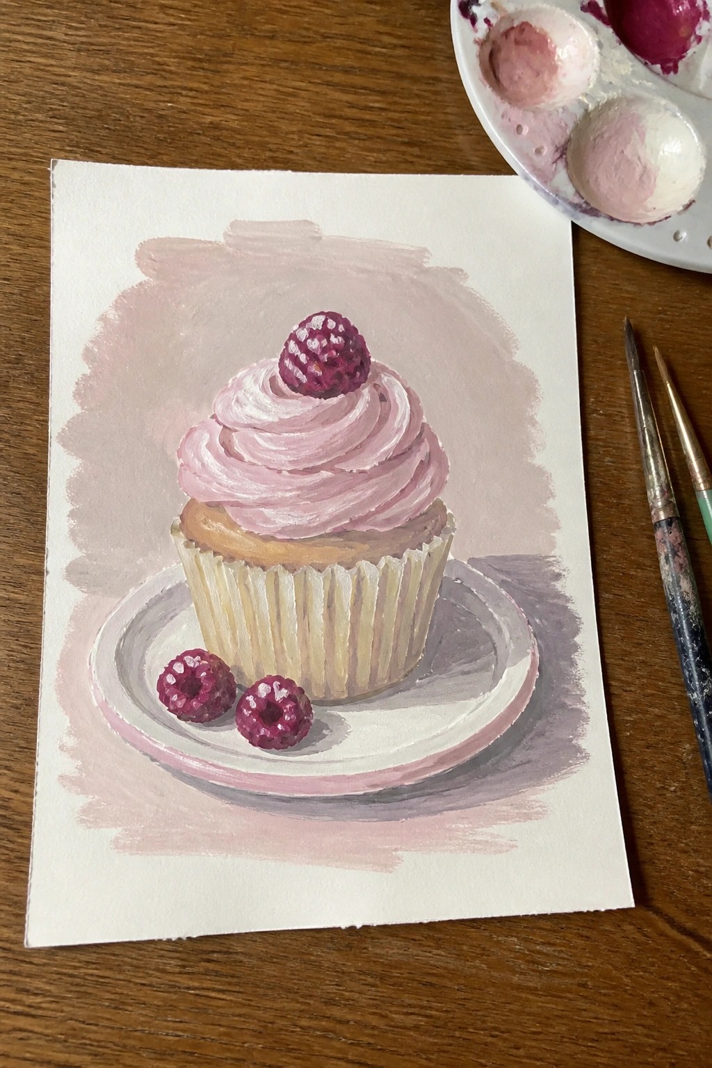 Watercolor of pink-frosted cupcake with raspberry on plate, palette and brushes nearby.