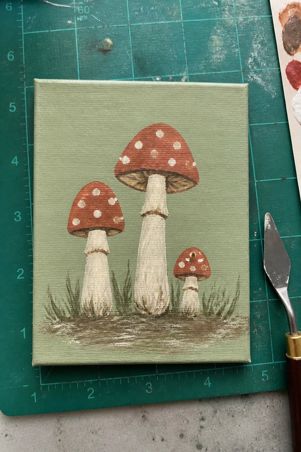 Acrylic painting of three mushrooms with red spotted caps on a light green background with grass at the bottom.