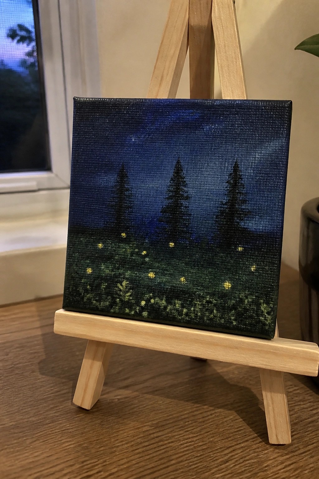 Small canvas on wooden easel shows starry blue night with three trees and glowing lights.