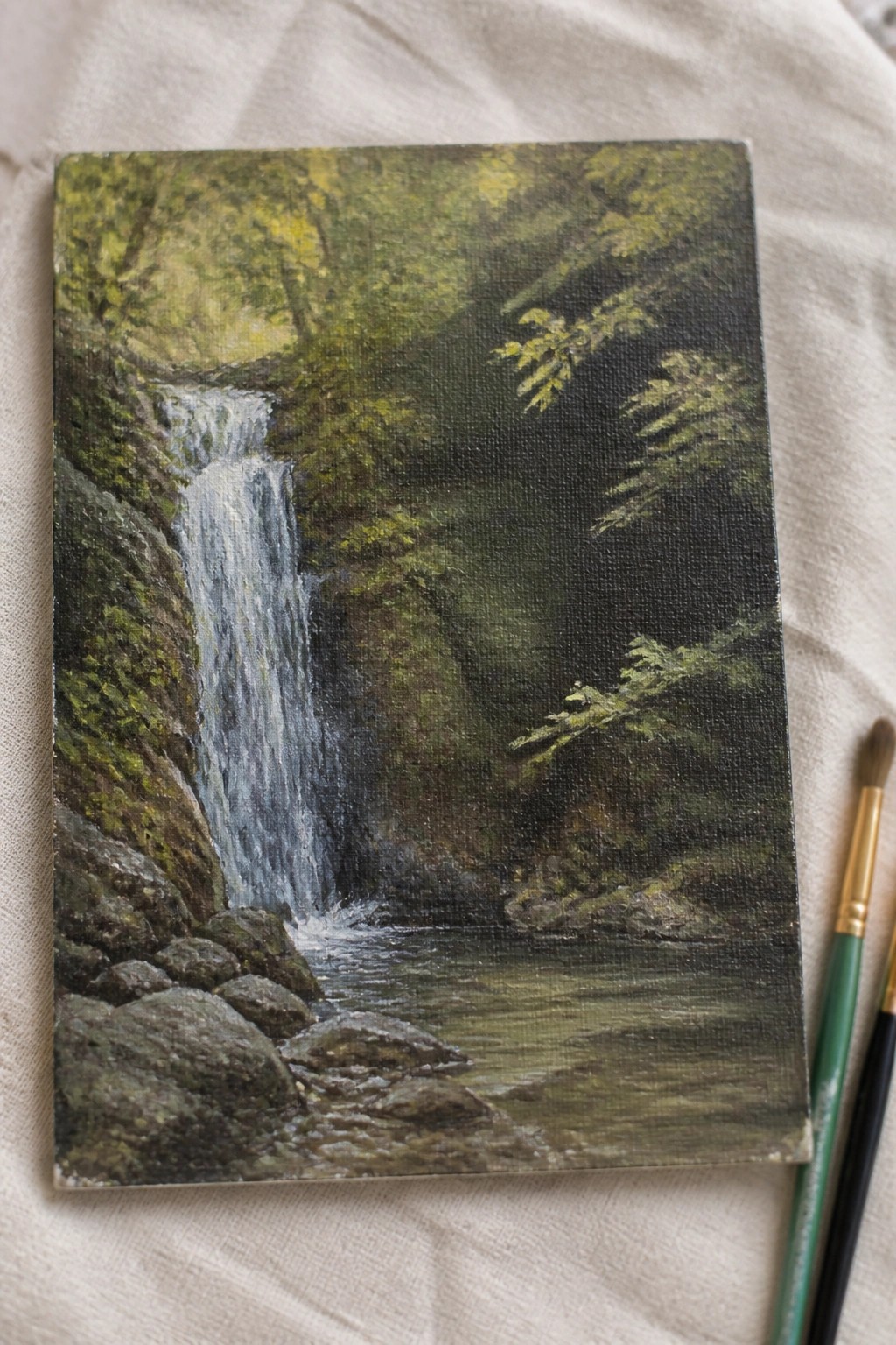 Small canvas painting of forest waterfall with rocks, pool, and two paintbrushes.