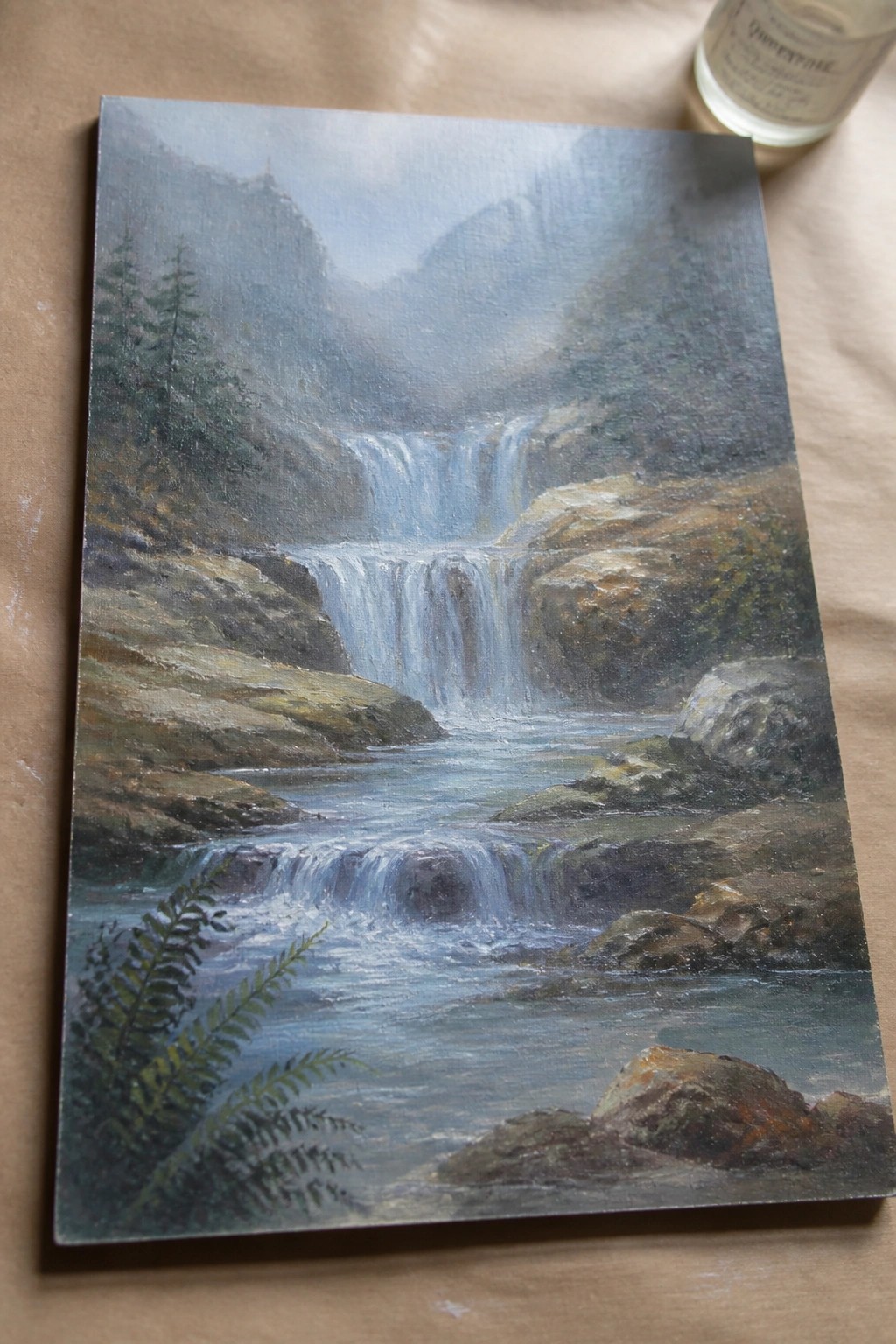 Oil painting of misty waterfall cascading over rocks in foggy mountains beside varnish bottle