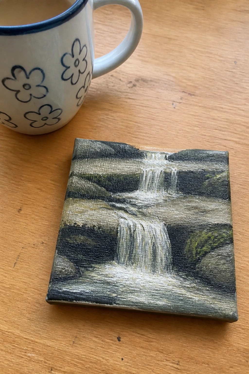 Small waterfall painting on canvas beside floral mug on wooden table.