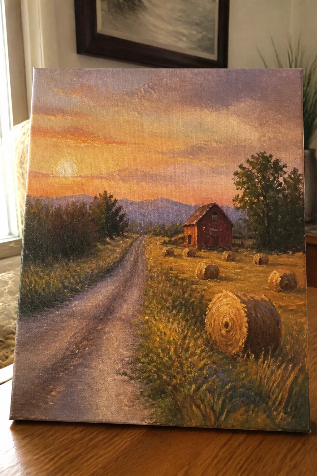 Rural sunset painting with red barn, hay bales, dirt road, and distant mountains