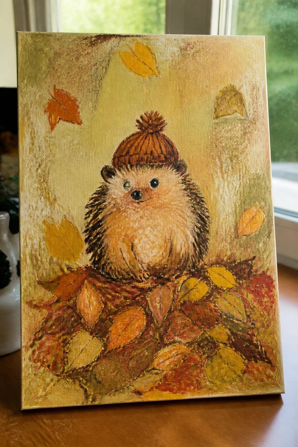 Hedgehog in brown knitted hat rests amid colorful autumn leaves on textured canvas.