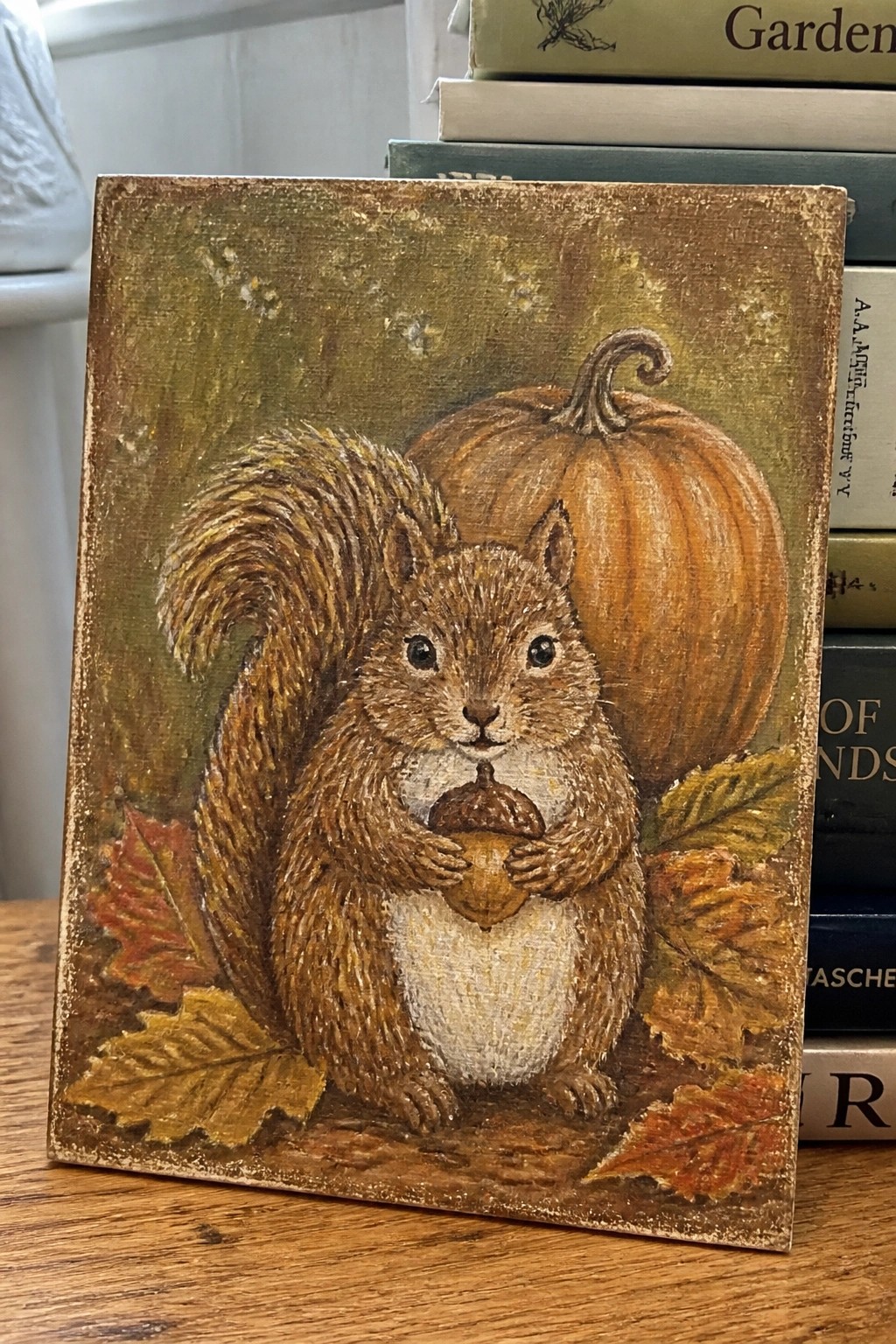 A painted canvas of a brown squirrel holding an acorn, with a pumpkin and autumn leaves behind it on a textured background.