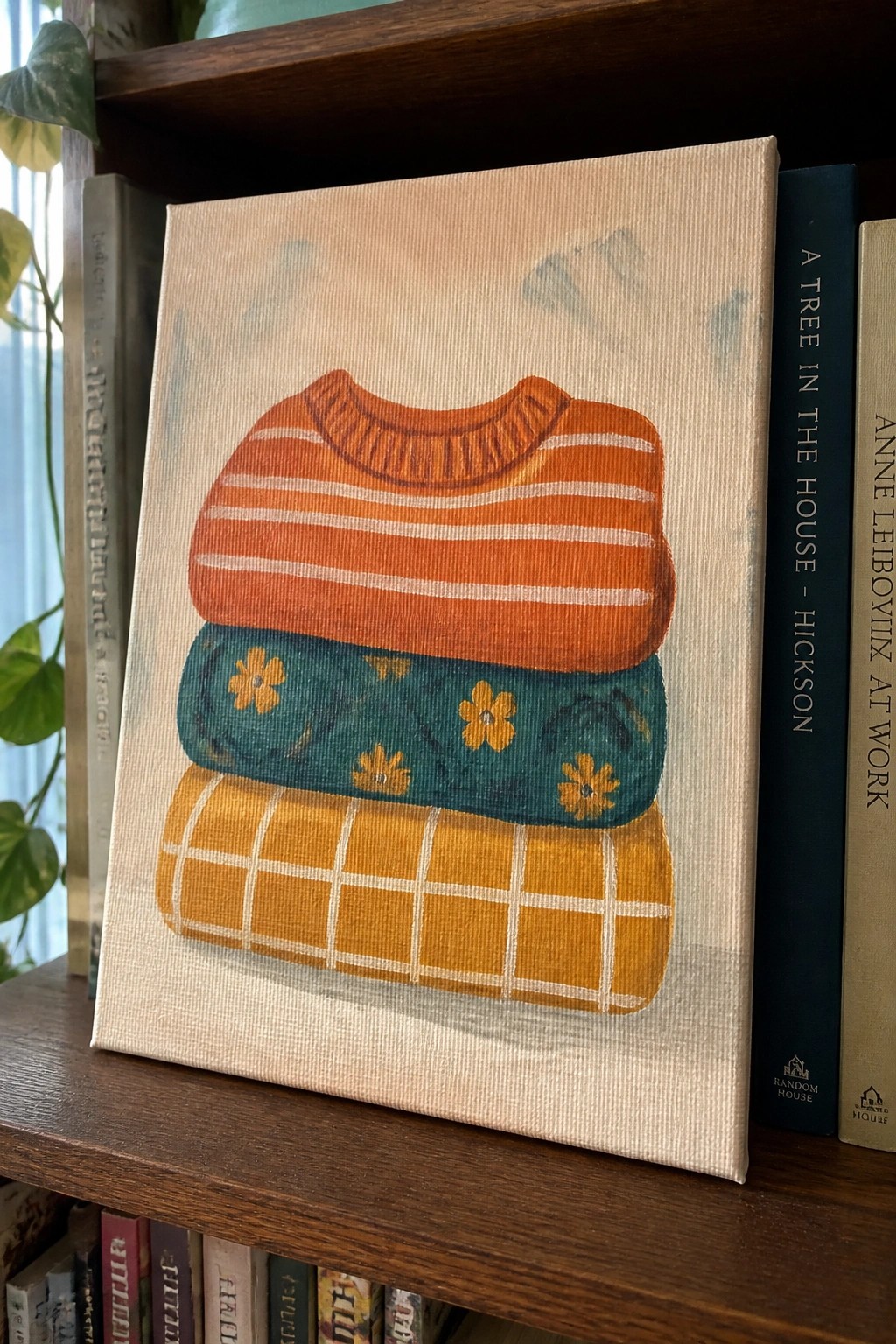A canvas painting of three stacked sweaters in orange stripes, teal with yellow flowers, and yellow plaid resting on a wooden bookshelf.