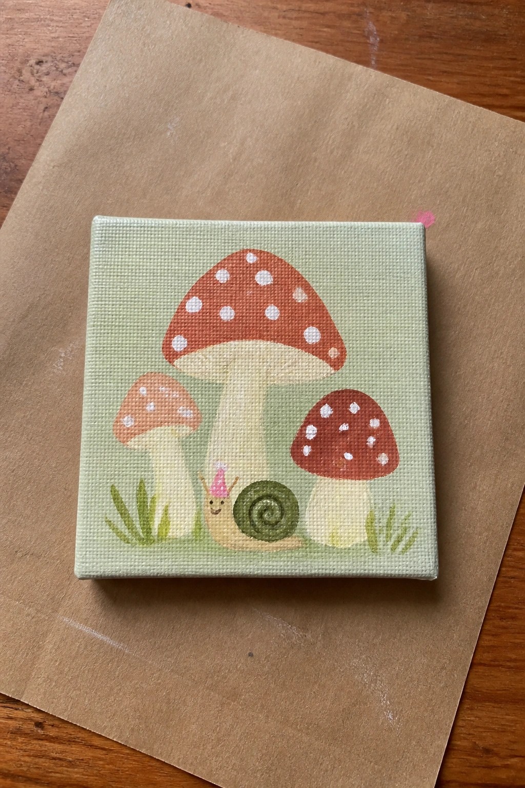Small square canvas painting of three red spotted mushrooms on a light green background with a snail wearing a pink party hat and green grass at the bottom.