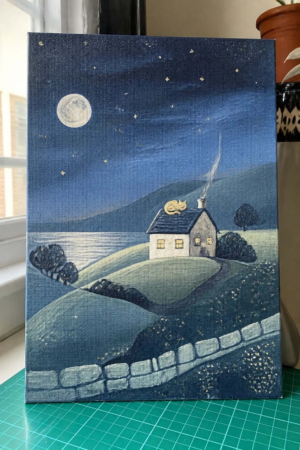 Whimsical painting of moonlit house with sleeping cat on roof amid starry hills.