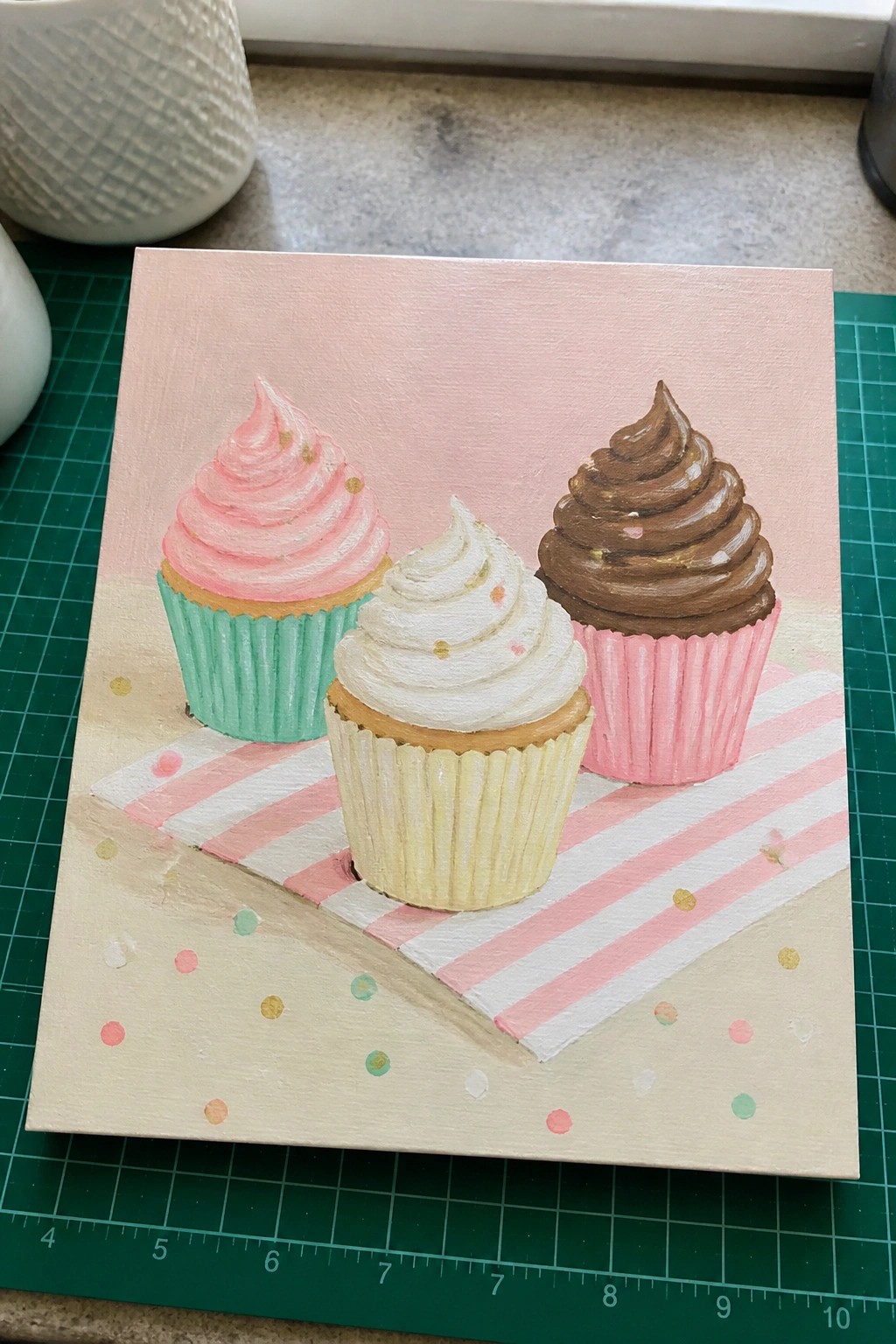 Acrylic painting of three cupcakes with pink, white, and brown frosting on striped paper.