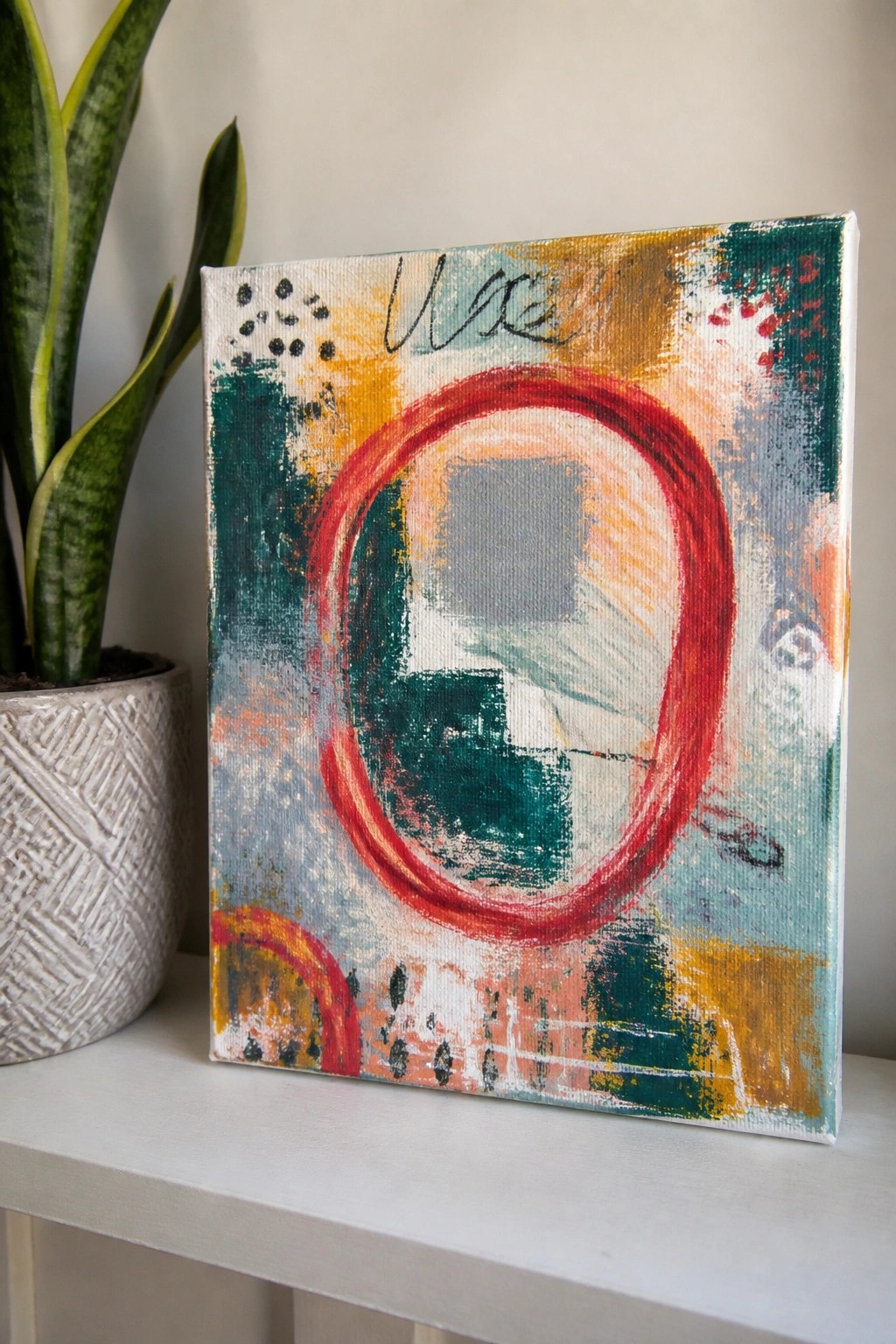 Abstract painting with bold red circle beside snake plant on white shelf.