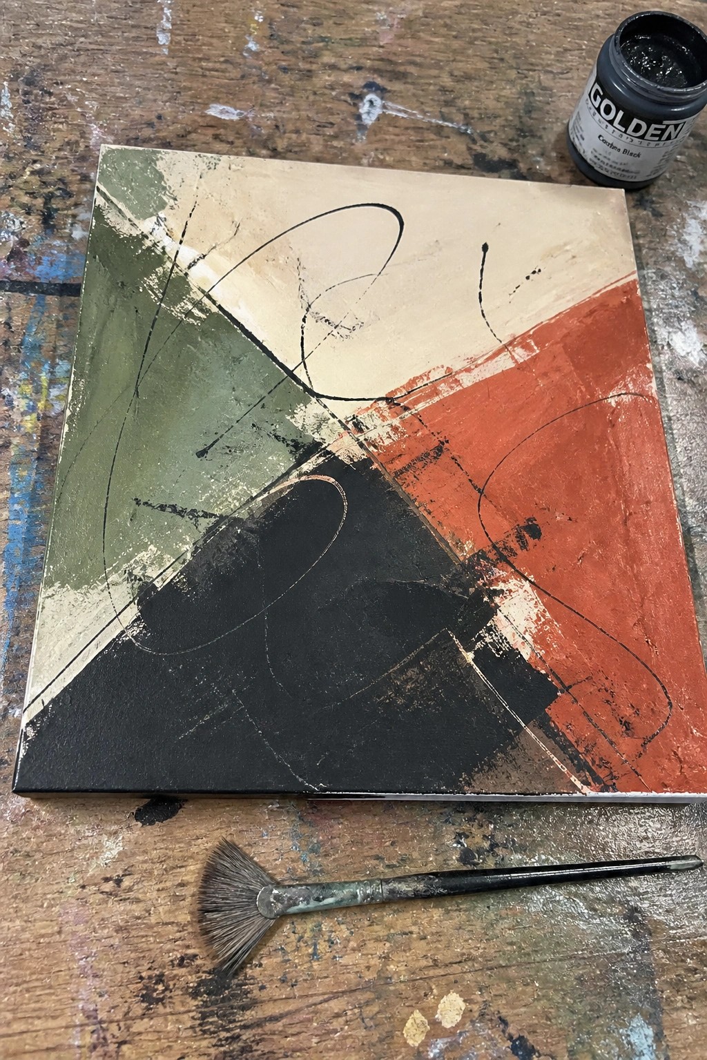 Abstract painting with green, black, red sections beside paint jar and fan brush on wood.