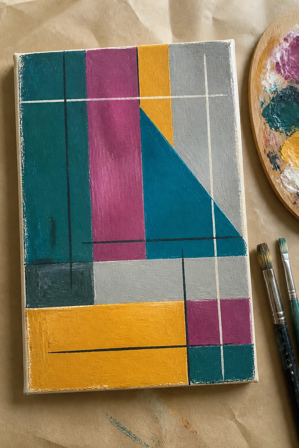 An abstract acrylic painting on canvas with teal, magenta, yellow, and gray geometric blocks divided by thin lines and a large teal triangle, shown next to brushes and a palette on brown paper.