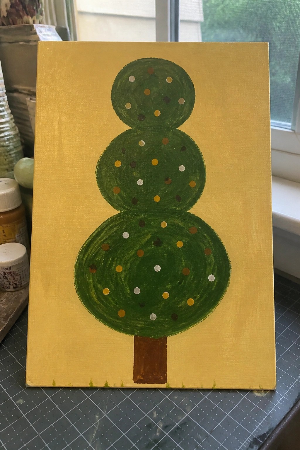 Three-tiered green abstract tree with colorful dots on yellow canvas with brown trunk.