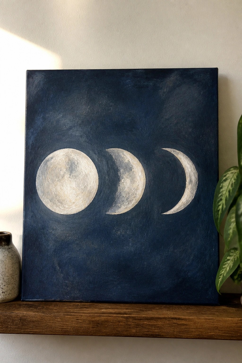 Three moon phases painted white on dark blue canvas atop wooden shelf.
