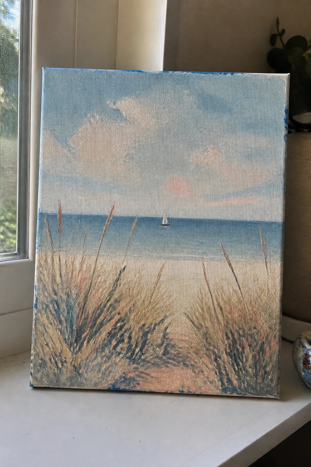 Impressionistic canvas painting of beach with tall grasses, ocean, sailboat under cloudy sky.