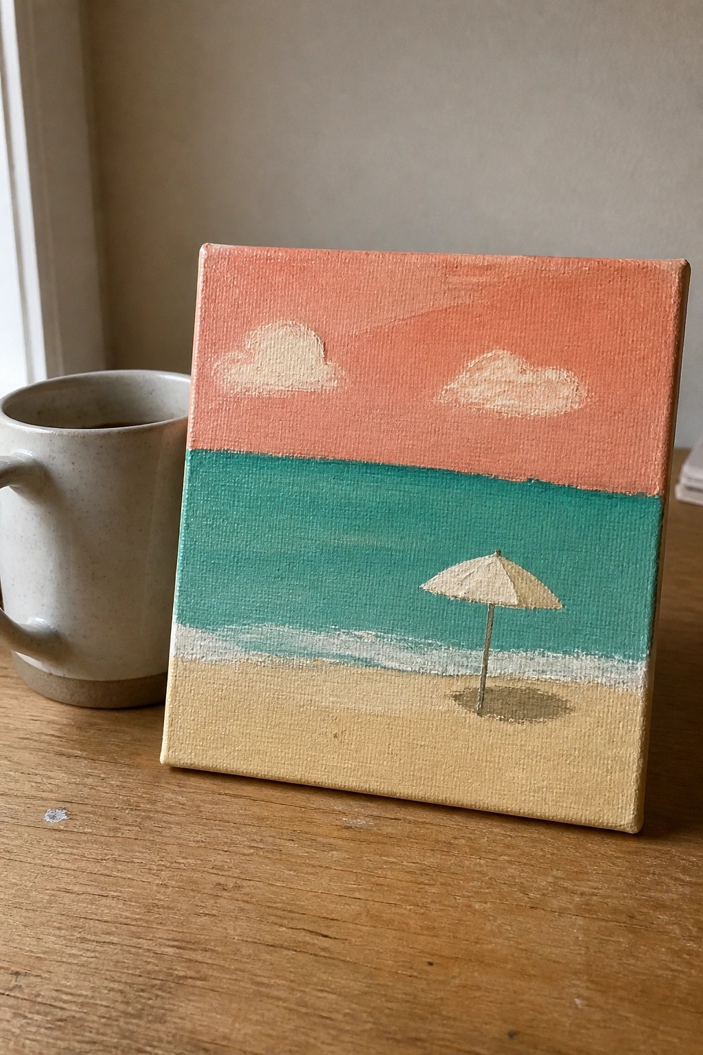 Small beach painting with peach sky, clouds, teal sea, umbrella beside mug on wood table