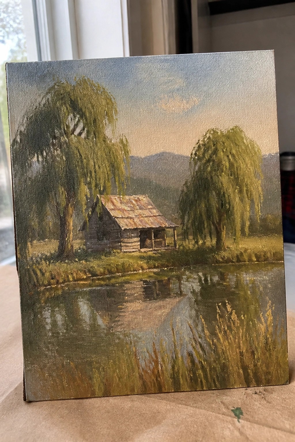 Log cabin with rusted roof by reflective pond, flanked by weeping willows and mountains.