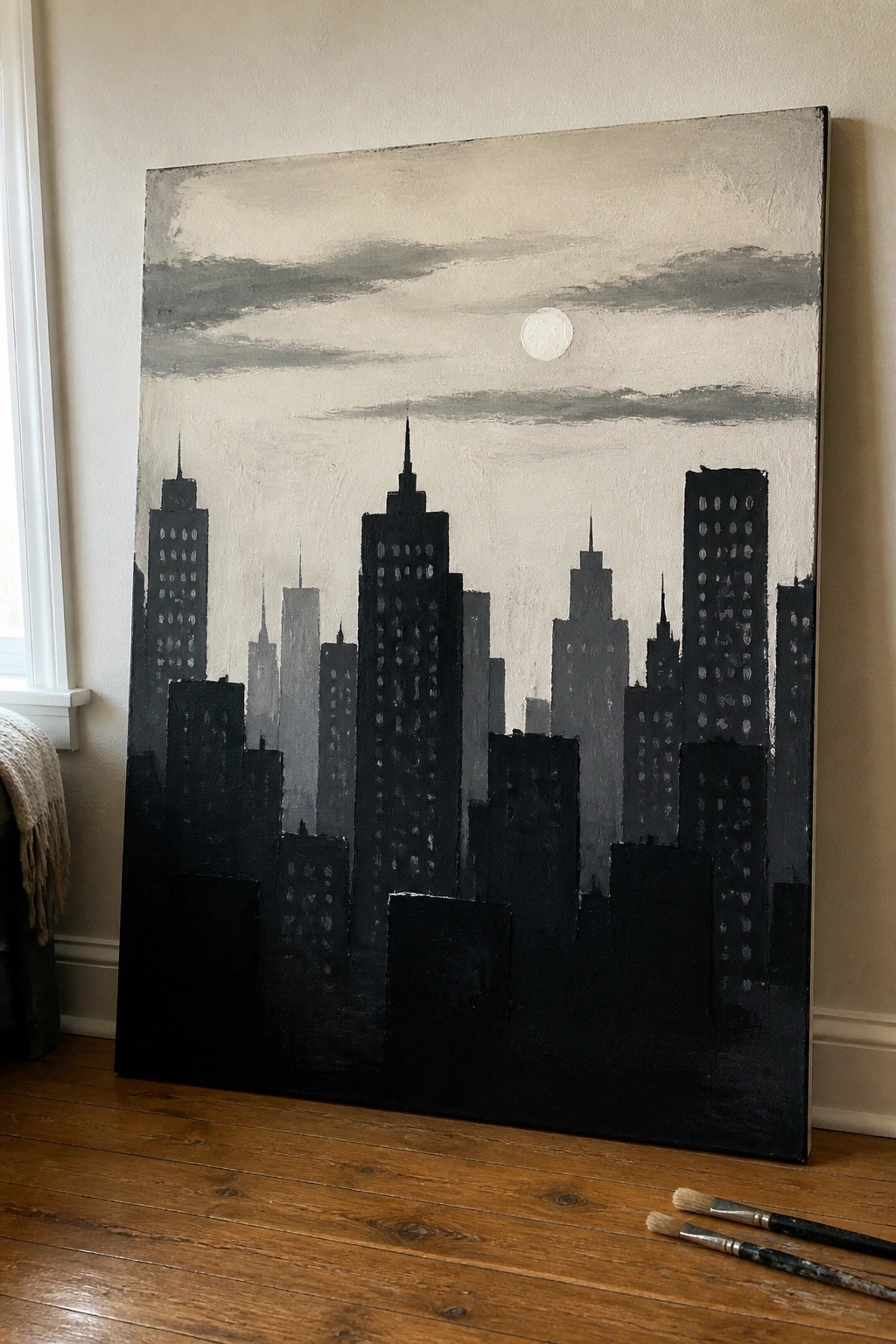 Large dark cityscape painting with skyscrapers and moon leaning on wooden floor beside brushes