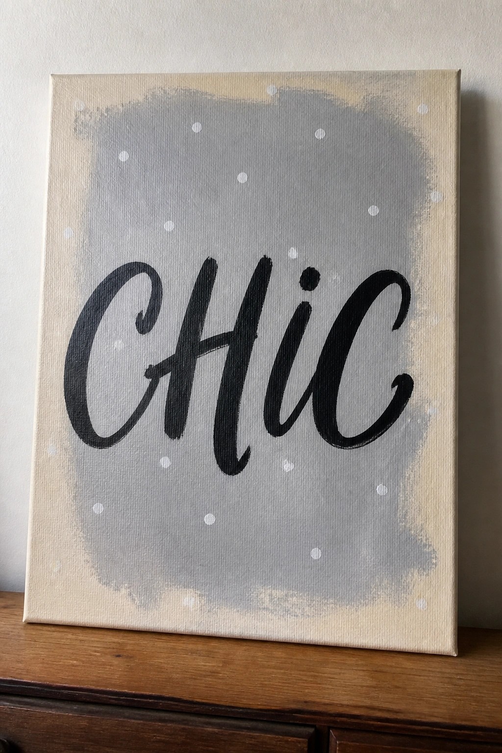 Canvas with black cursive 'CHiC' on gray dotted beige background atop wood.