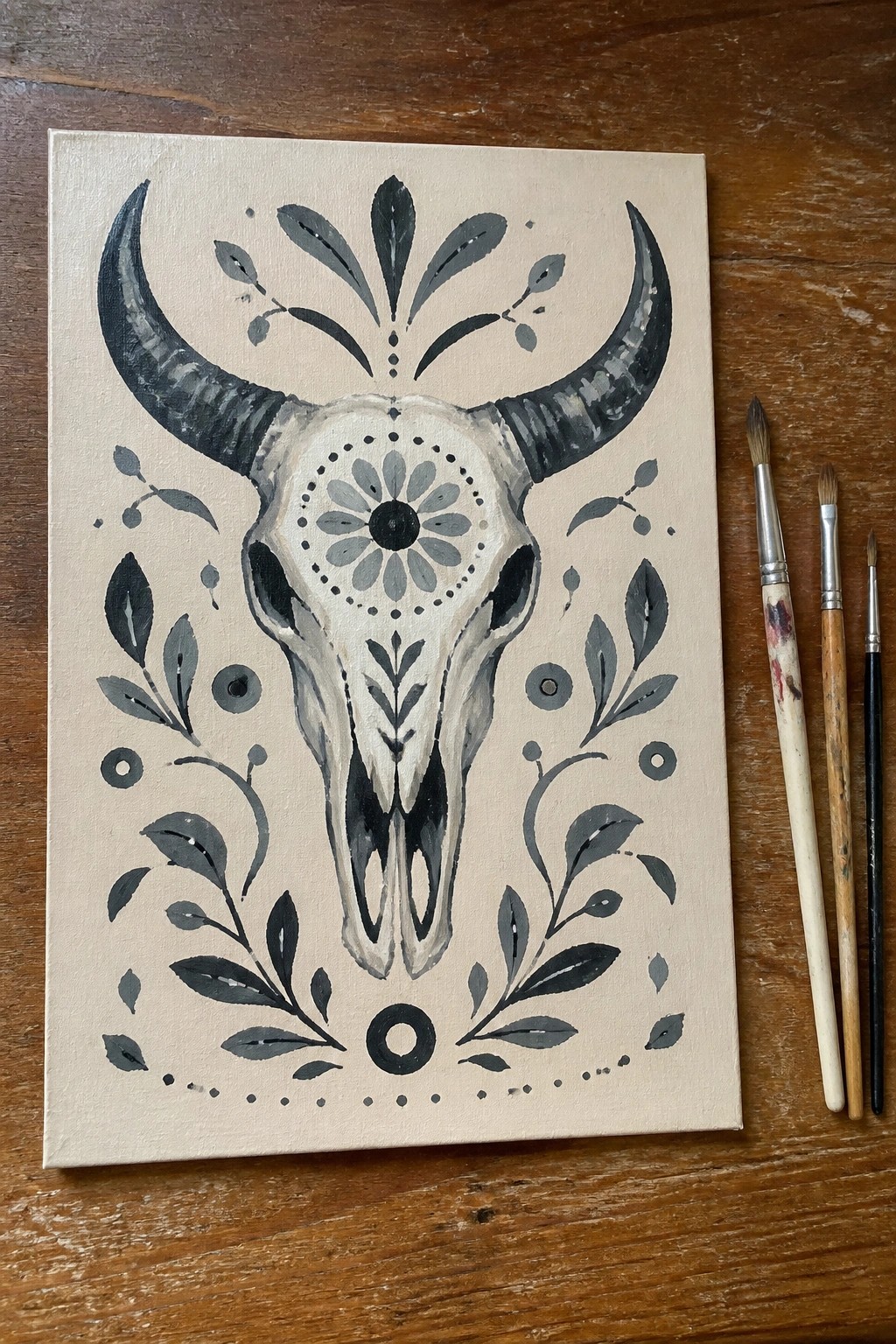 Ornate bull skull painting with horns and floral designs beside paintbrushes on wood