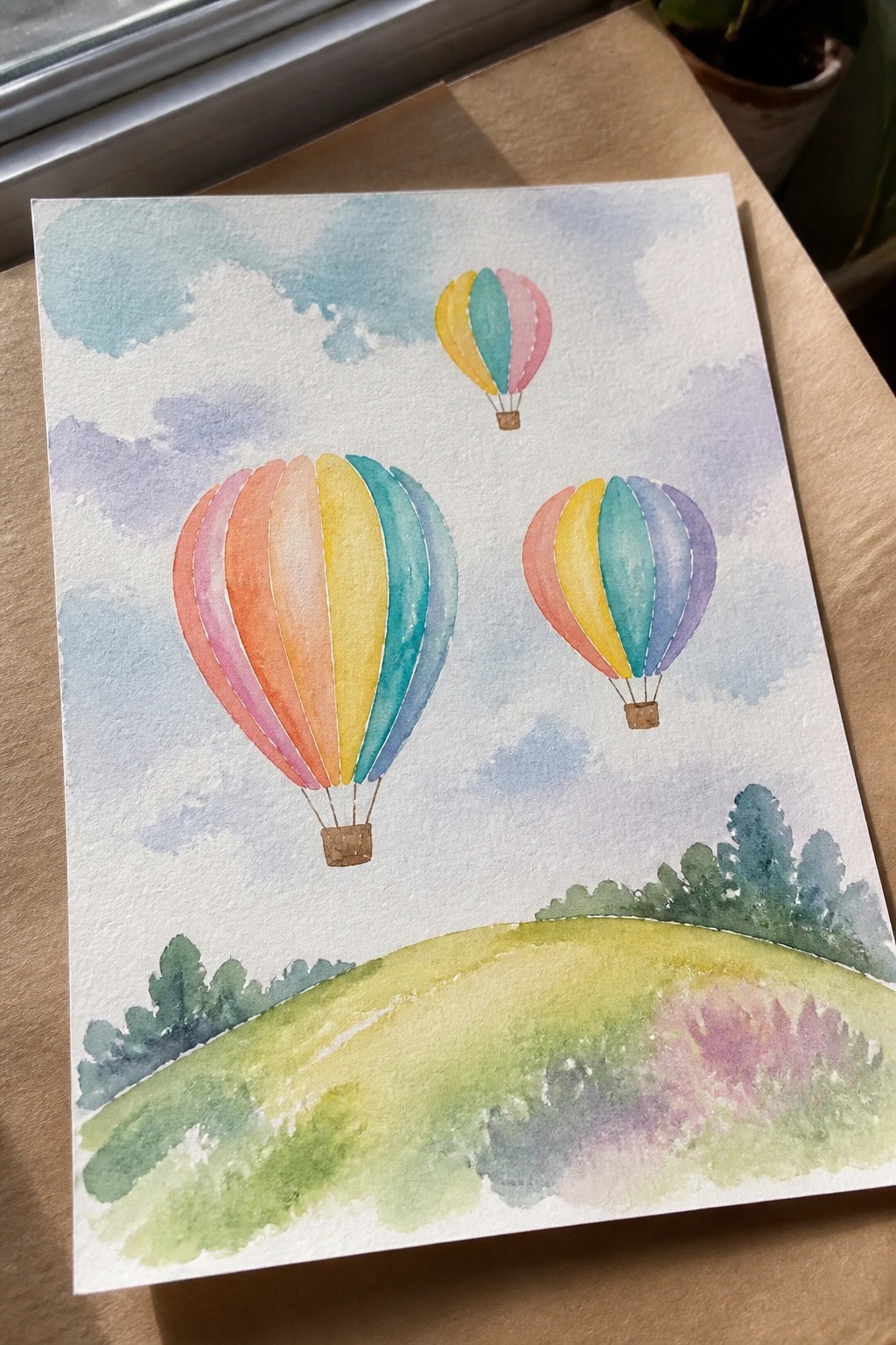 Watercolor painting of three colorful hot air balloons above a green hill with trees.